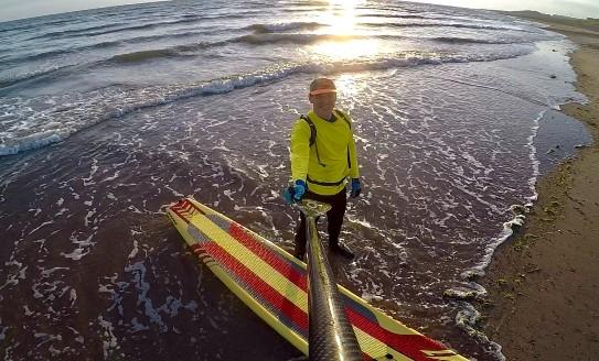 Adrian taking a selfie with a selfie stick, standing on his paddle board at the water's edge on a beach