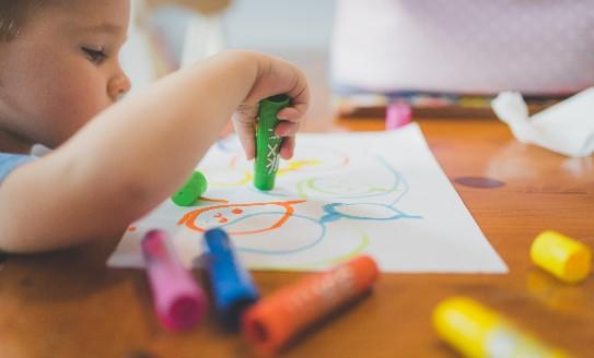 A young boy sitting at a table with different coloured crayons, drawing on a piece of paper