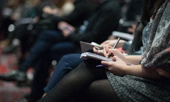 Conference attendees sitting in a line, woman in foreground taking notes in notebook 