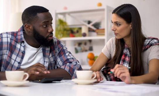 A Black man and a Caucasian woman sit at the table with two cups of tea, going through a budget sheet with a pencil, looking concerned