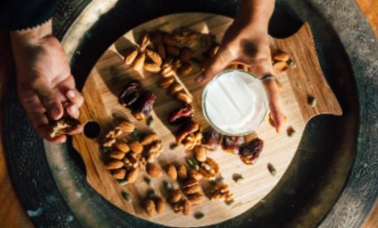 Two hands over a wooden cutting board, one holding a glass of milk and the other holding a date. There are dates and nuts on the cutting board. 