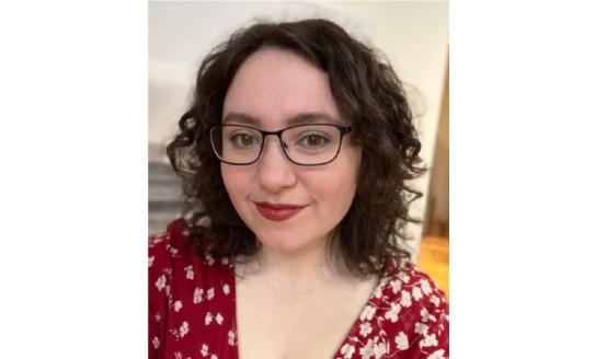 Holly smiling to camera. She has short, dark curly hair, glasses and she's wearing a red and white spotty dress