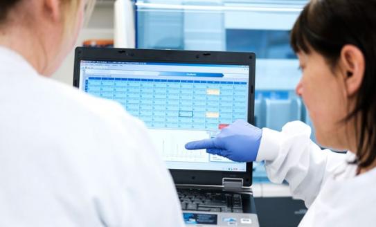 Two scientists in white lab coats with their backs to camera, looking at a computer screen together