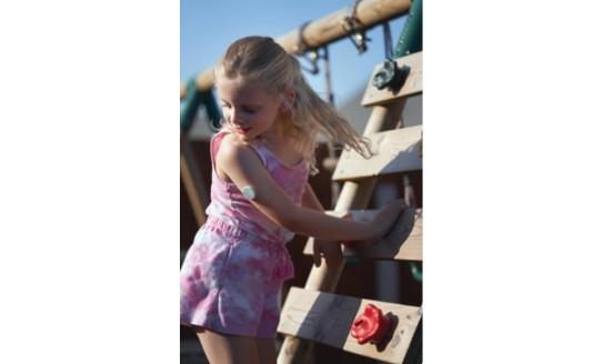 A photo of Libby, a young girl with blonde hair wearing a pink top and shorts, playing on a climbing frame. You can see her continuous glucose monitor on her arm.