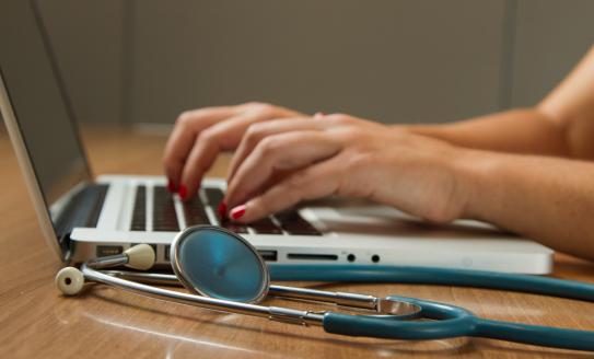 A pair of hands wearing nail red varnish typing on a laptop with a stethoscope laying on the desk next to it