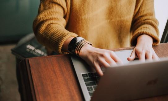 A person in a yellow jumper at a desk, typing on a laptop