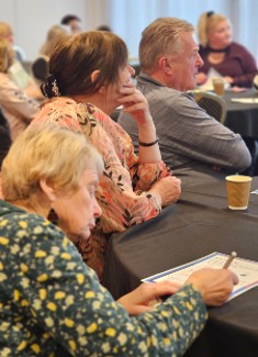 Three people sit round a table looking straight ahead whilst one woman takes notes