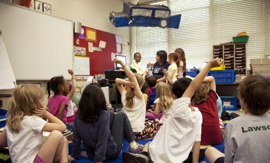 Primary school children sitting on the carpet in front of a whiteboard as their teacher reads to them