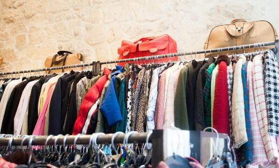 A rack of secondhand clothes in a shop, with handbags on top of the rack, against a white wall