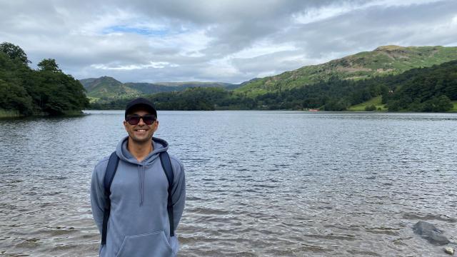 A man wearing sunglasses stands by a lake, smiling