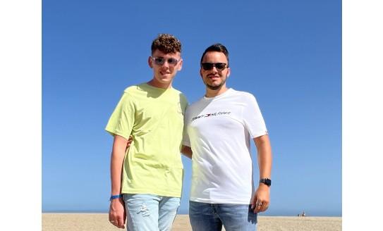 Two young men, one wearing a yellow tshirt and one wearing a white tshirt, standing next to eachother on a beach and smiling to camera